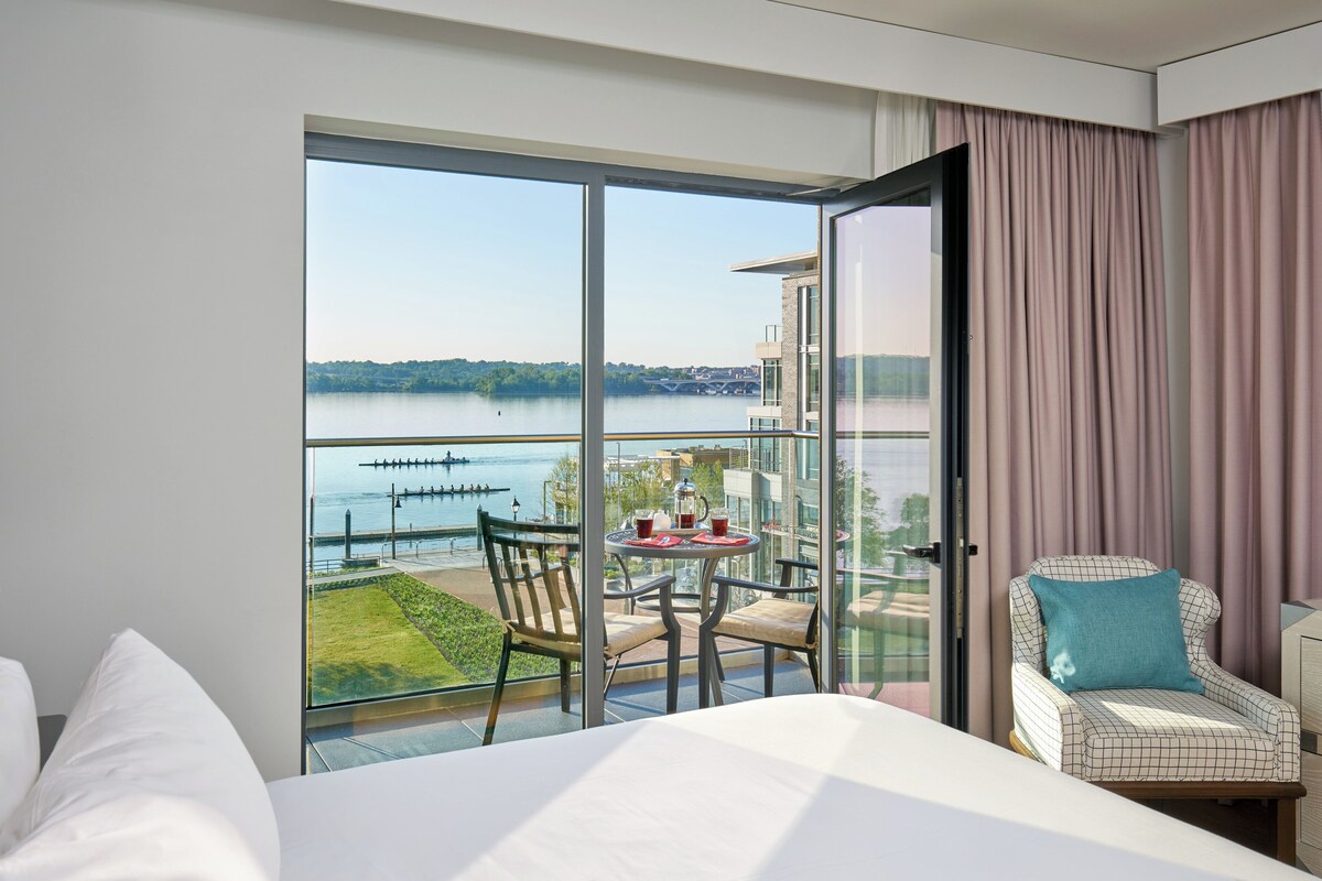 Une chambre d’hôtel dotée d’une grande porte-fenêtre coulissante donnant sur un balcon avec vue sur un lac et un pont. On y trouve un lit avec du linge de lit blanc, une petite table ronde recouverte d’une nappe rouge et une chaise avec un coussin bleu.