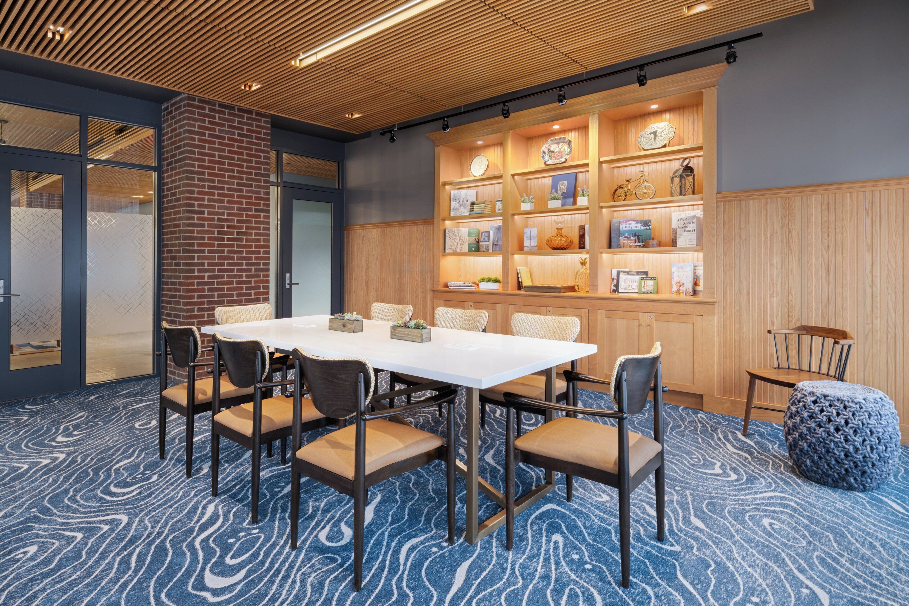 A conference room with a white table, six chairs, a brick wall, and a wooden panel wall with shelves.