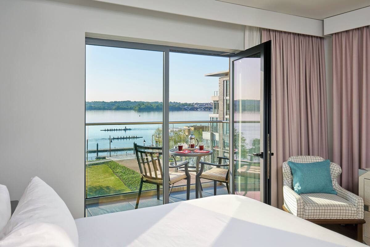 Une chambre d’hôtel dotée d’une grande porte-fenêtre coulissante donnant sur un balcon avec vue sur un lac et un pont. On y trouve un lit avec du linge de lit blanc, une petite table ronde recouverte d’une nappe rouge et une chaise avec un coussin bleu.