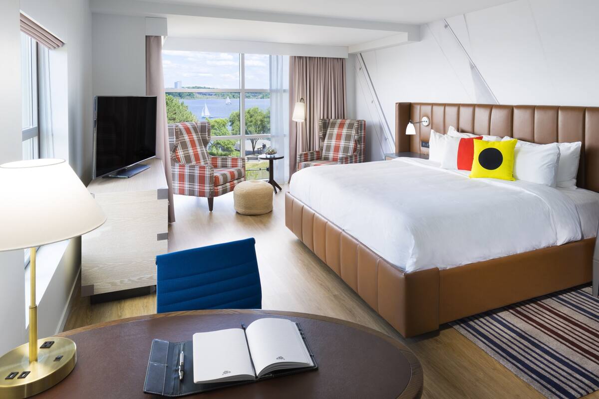 Ett hotellrum med en stor säng, ett skrivbord med en lampa, en TV, två stolar, ett litet bord och utsikt över omgivningarna.