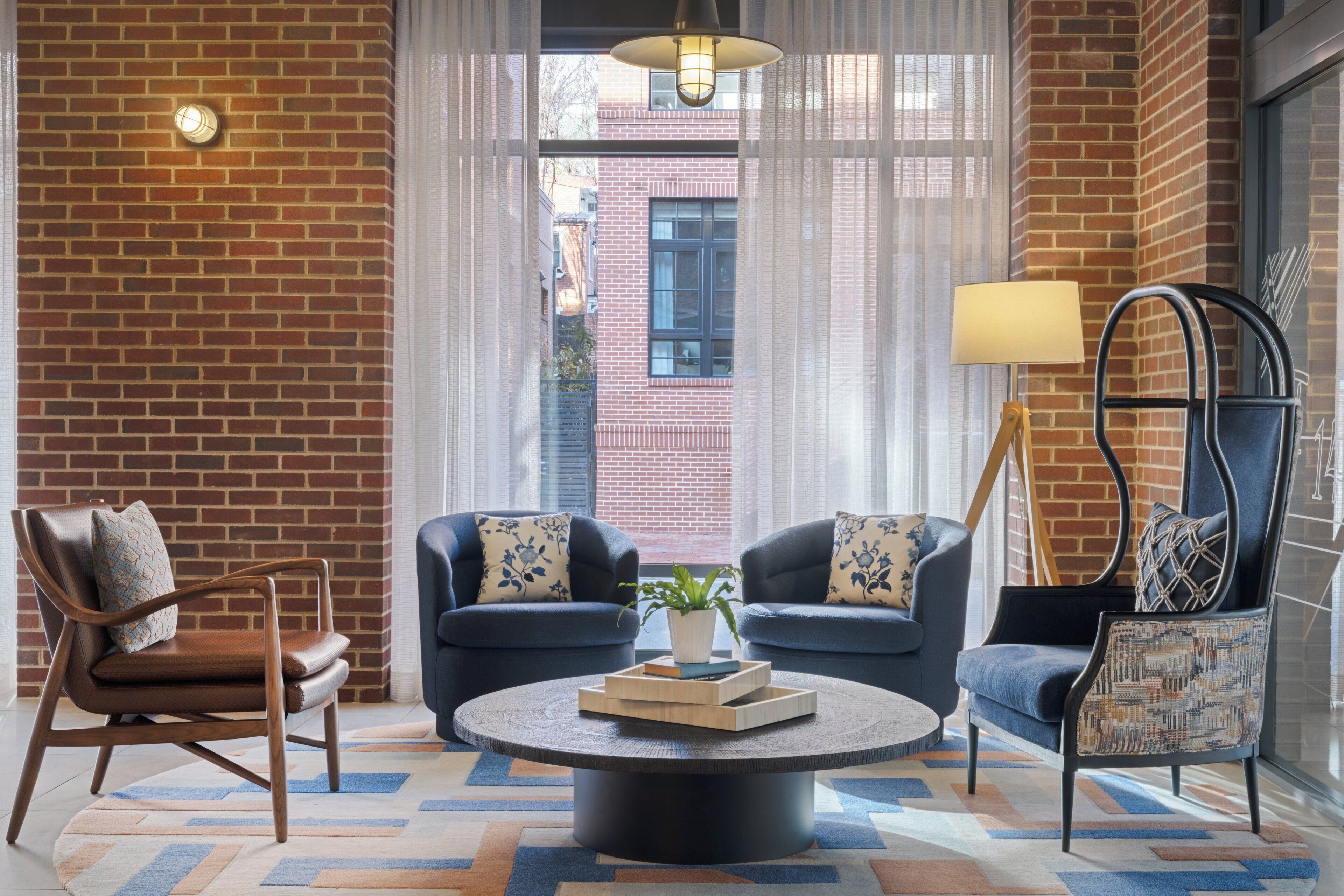 A room with a brick wall, two sofas with patterned cushions, a wooden chair, a round coffee table with a plant and books, and a lamp.