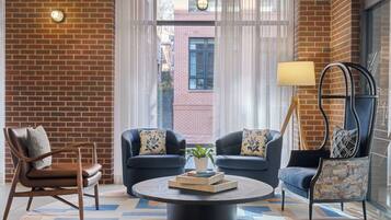 A room with a brick wall, two sofas with patterned cushions, a wooden chair, a round coffee table with a plant and books, and a lamp.