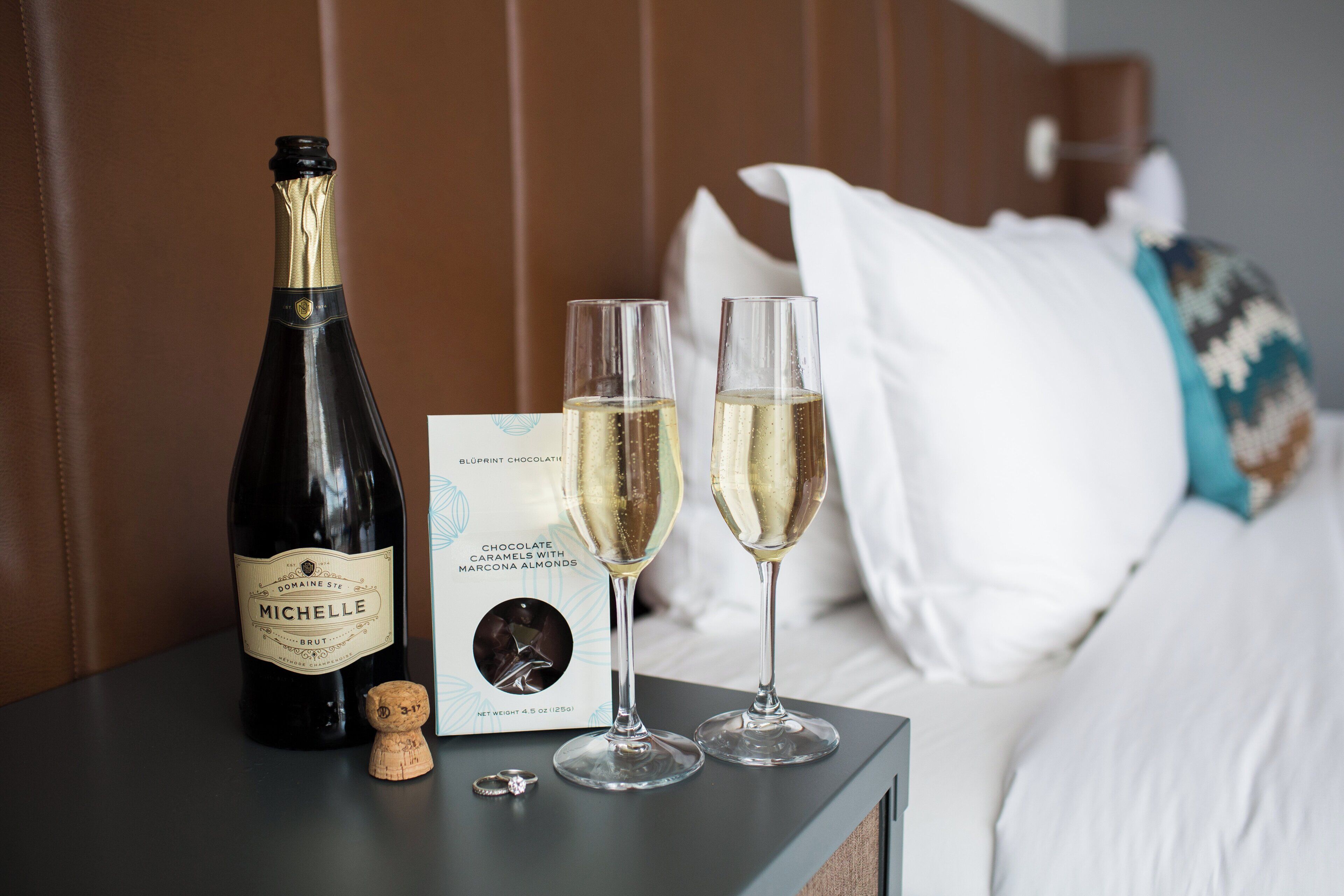 A bottle of champagne, two glasses, and a menu on a bedside table in a hotel room.