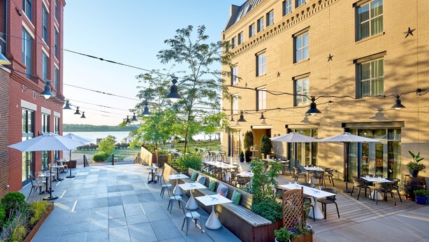 Eine Außenpatio mit Tischen und Stühlen, umgeben von Ziegelgebäuden, mit Blick auf ein Gewässer.