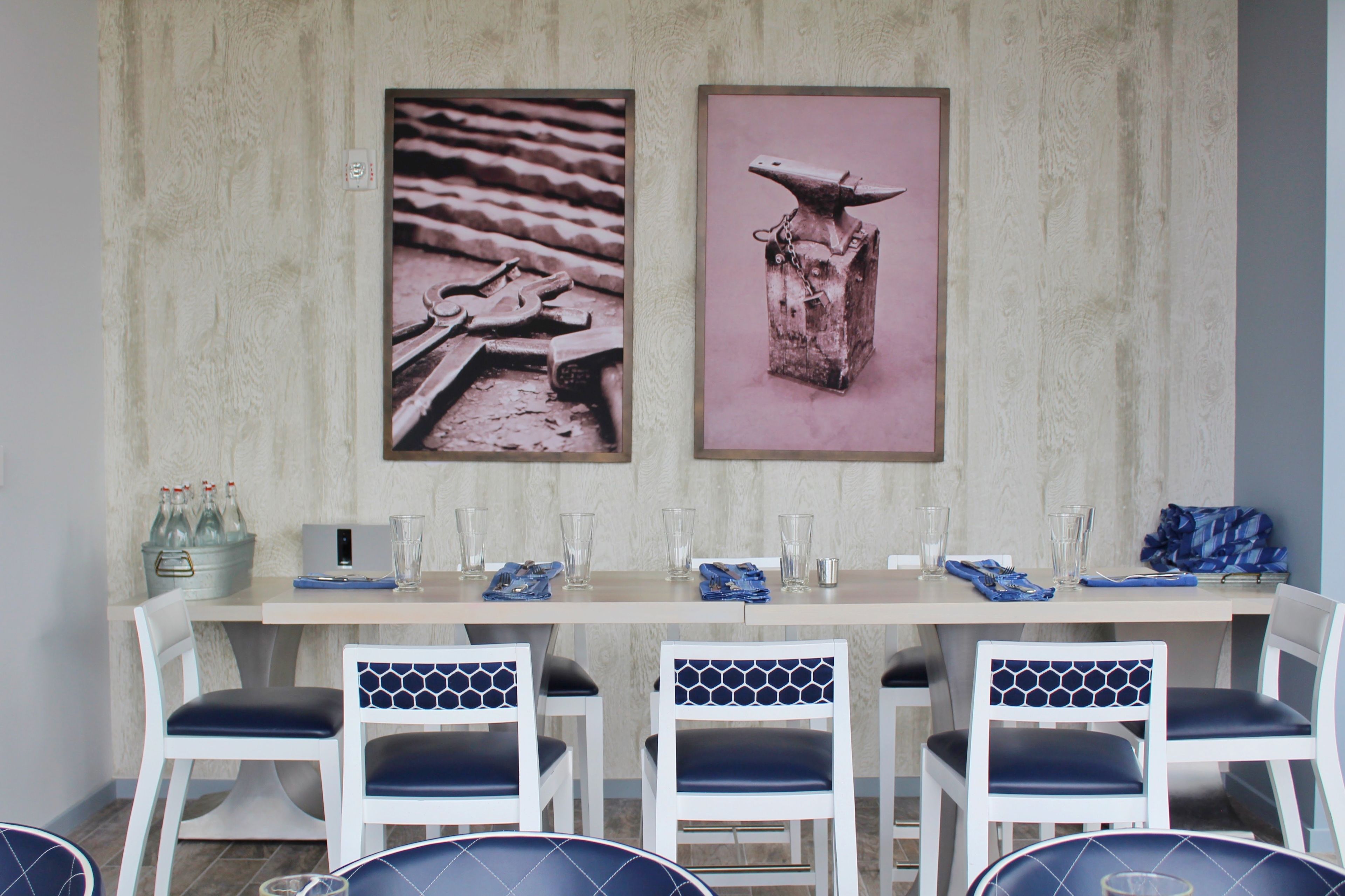 A dining area with a long table, white chairs with blue cushions, and two framed pictures on the wall.