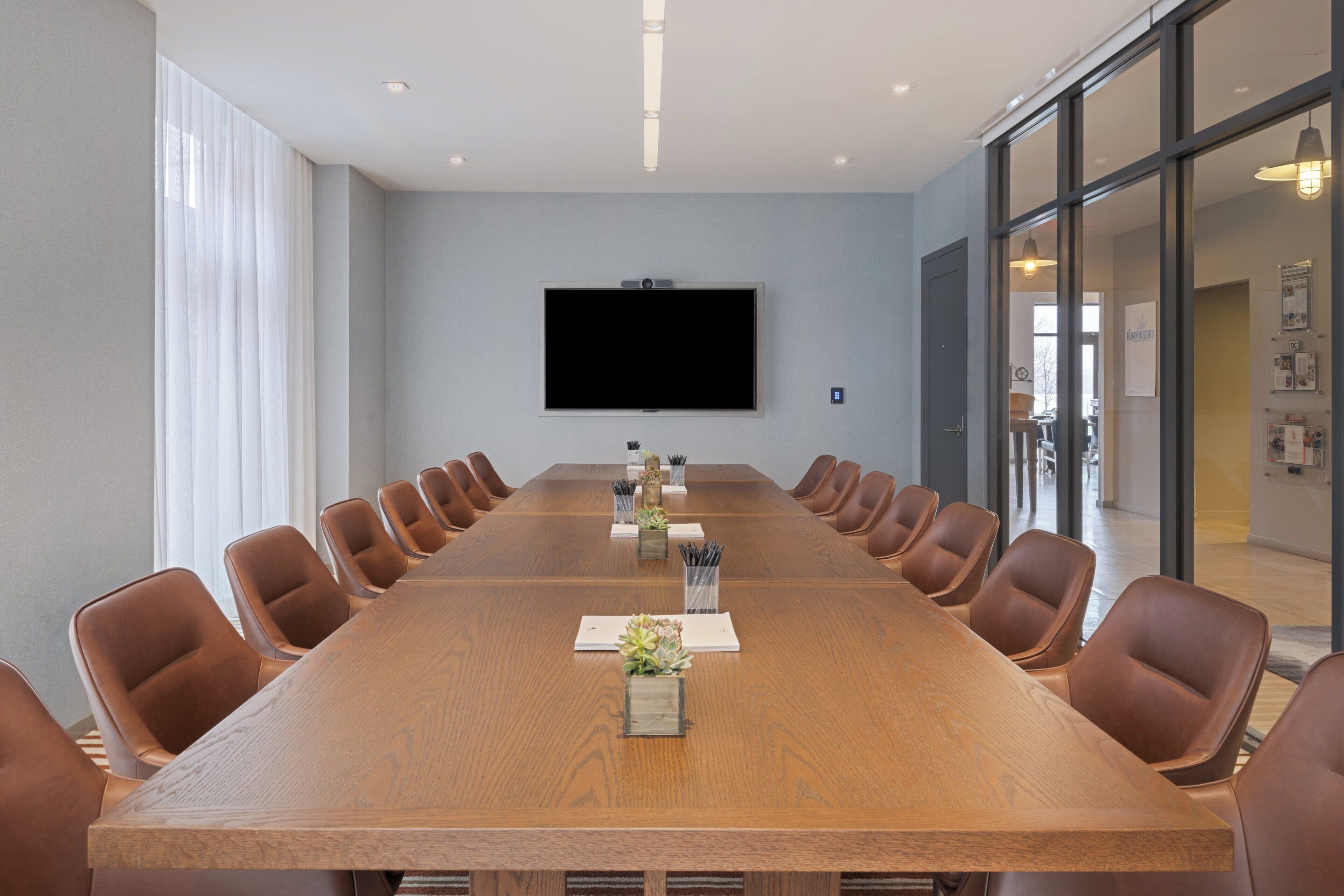 A conference room with a long table, chairs, a flat-screen TV, and a glass wall.
