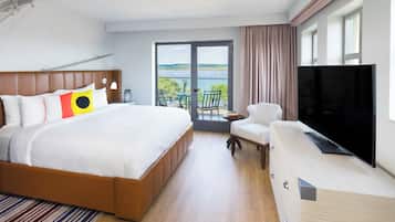 Ein Hotelzimmer mit einem großen Bett, einem Fernseher, einem Balkon mit Blick auf das Wasser und einem Sessel.