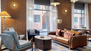 Ein modernes Wohnzimmer mit Backsteinwand, großen Fenstern, einem braunen Ledersofa, einem blauen Sessel, einem runden Holztisch und einem Essbereich im Hintergrund.