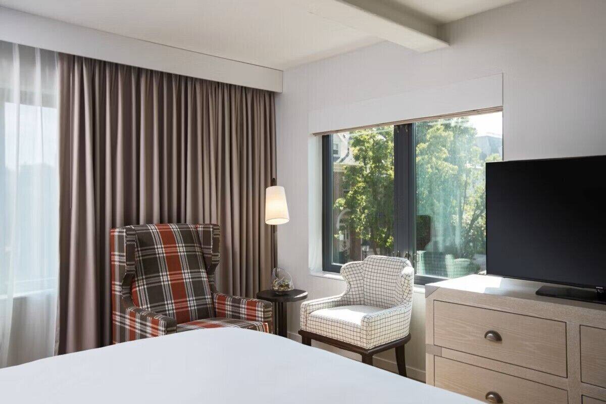 Ett hotellrum med en säng, två fåtöljer med rutigt tyg, en tv, en lampa och ett fönster med utsikt över träd.