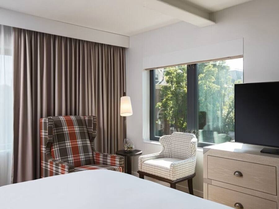 A hotel room with a bed, two plaid chairs, a TV, a lamp, and a window with a view of trees.
