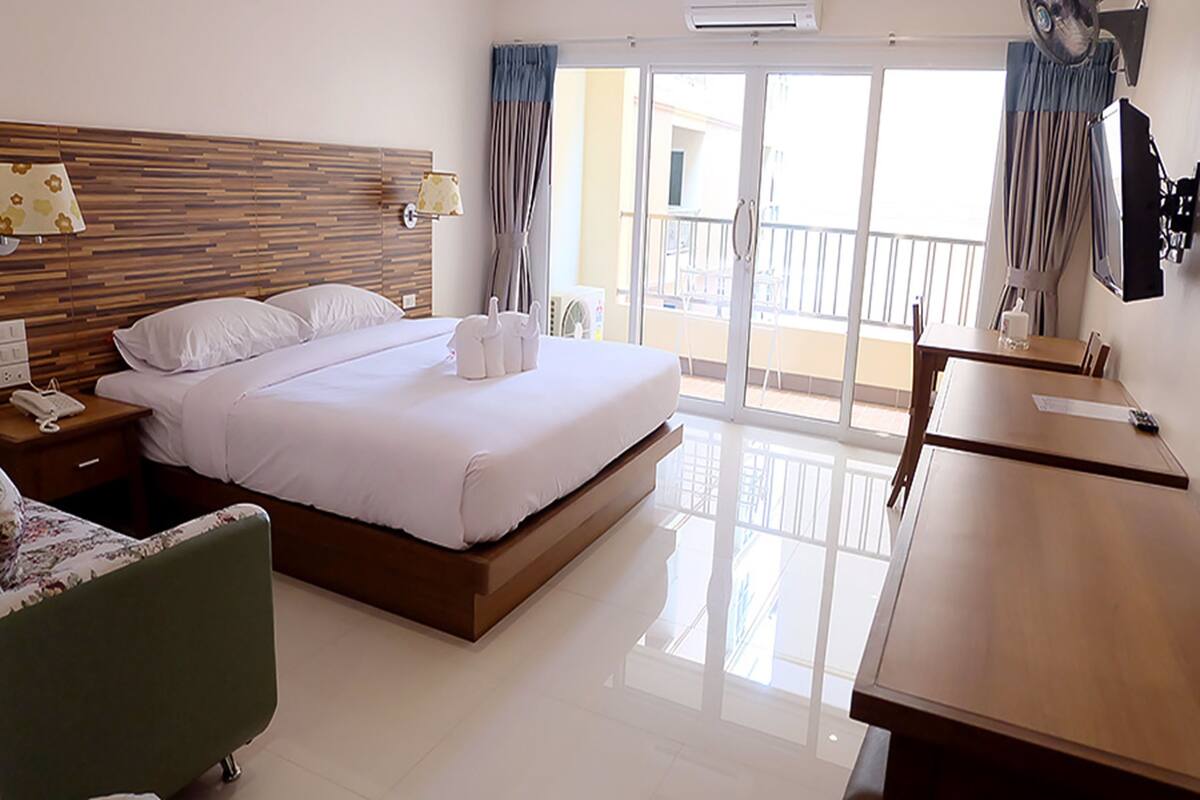 A hotel room with a large bed, a bedside table with a lamp, a chair, a desk, and a balcony with a view.