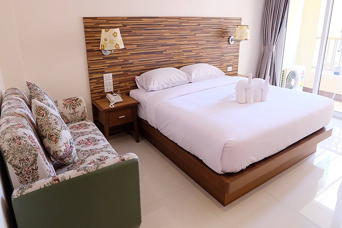 A modern hotel room with a large bed, a wooden headboard, a bedside table with a phone, and a green chair.