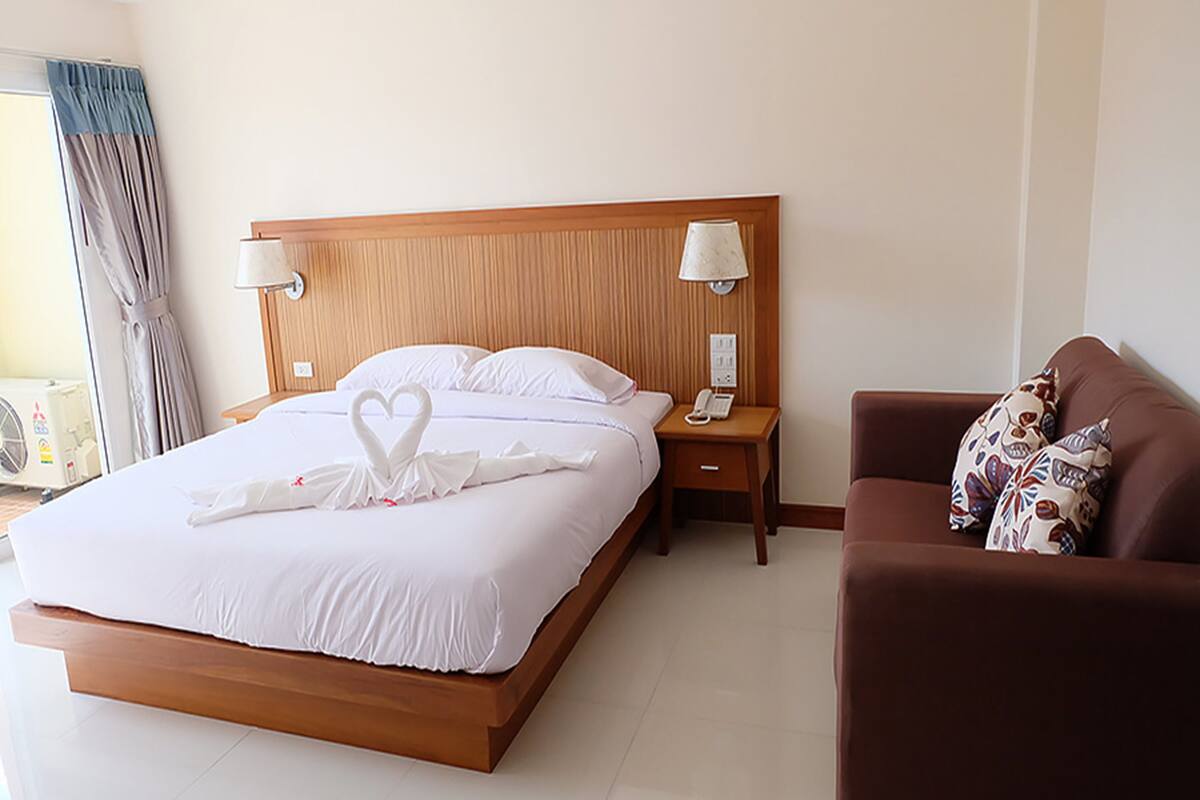 A hotel room with a wooden bed, white bedding, a brown sofa, and a bedside table with a lamp.