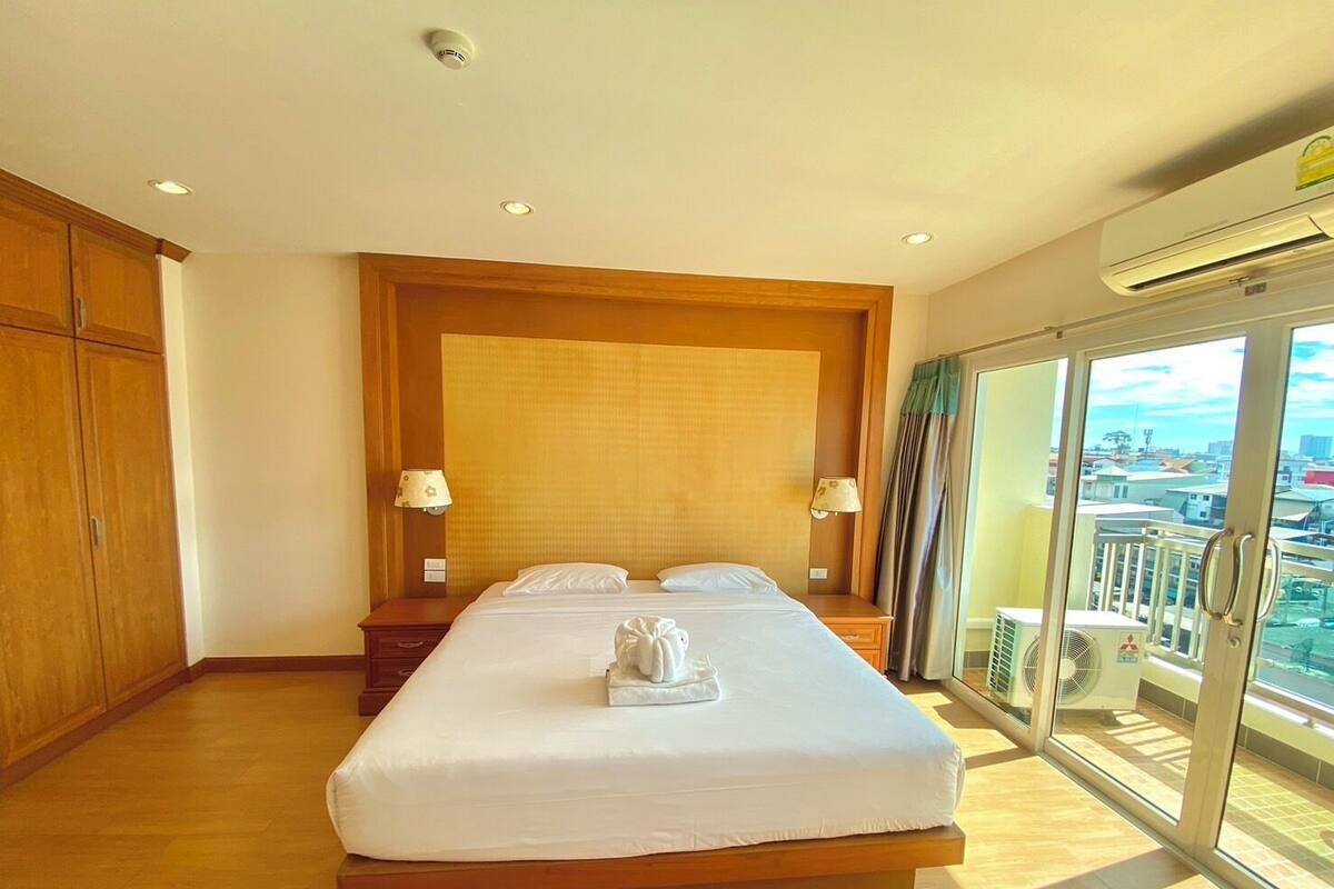 A hotel room with a large bed, wooden headboard, two bedside lamps, a window with a view of the city, and an air conditioning unit.