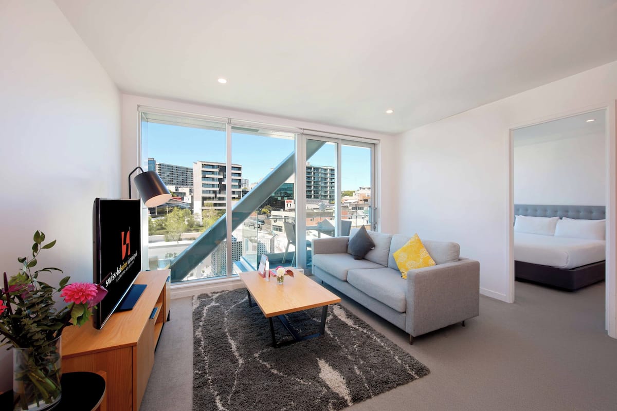Ein modernes Wohnzimmer mit Sofa, Couchtisch und Fernseher. Durch das große Fenster ist eine Treppe zu sehen, und im Hintergrund befindet sich ein Bett.