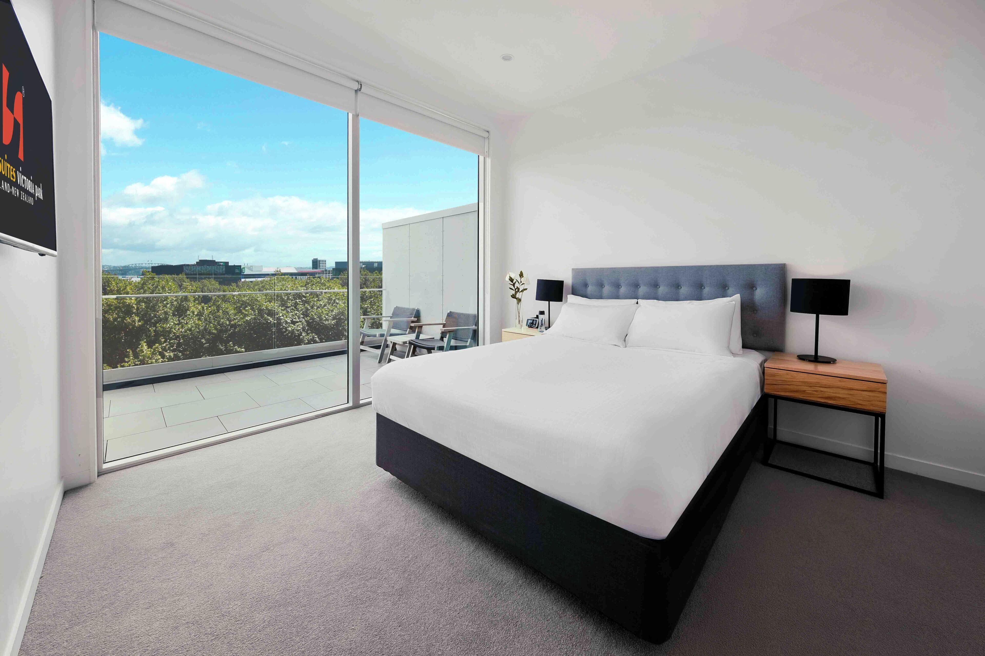 A modern bedroom with a large bed, a nightstand, and a view of a balcony with a cityscape.