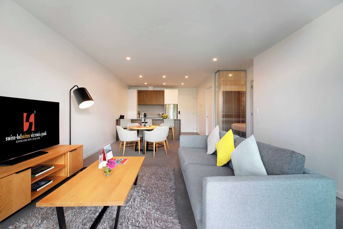 Ein modernes Hotelzimmer mit grauem Sofa, einem Holztisch, einem Fernseher und einem Essbereich mit weißem Tisch und Stühlen.