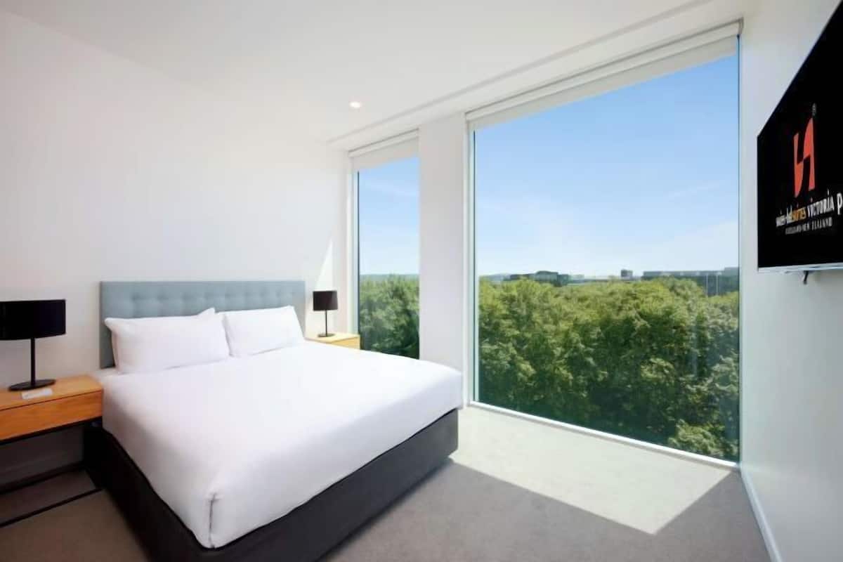 Ein modernes Hotelzimmer mit einem großen Bett, einem Nachttisch, einer Lampe und einem großen Fenster mit Blick auf Bäume.