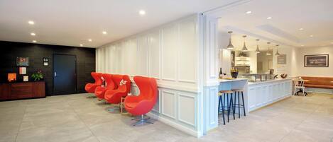 A modern hotel lobby with a reception desk, red chairs, a bar area, and a seating area with a bench.