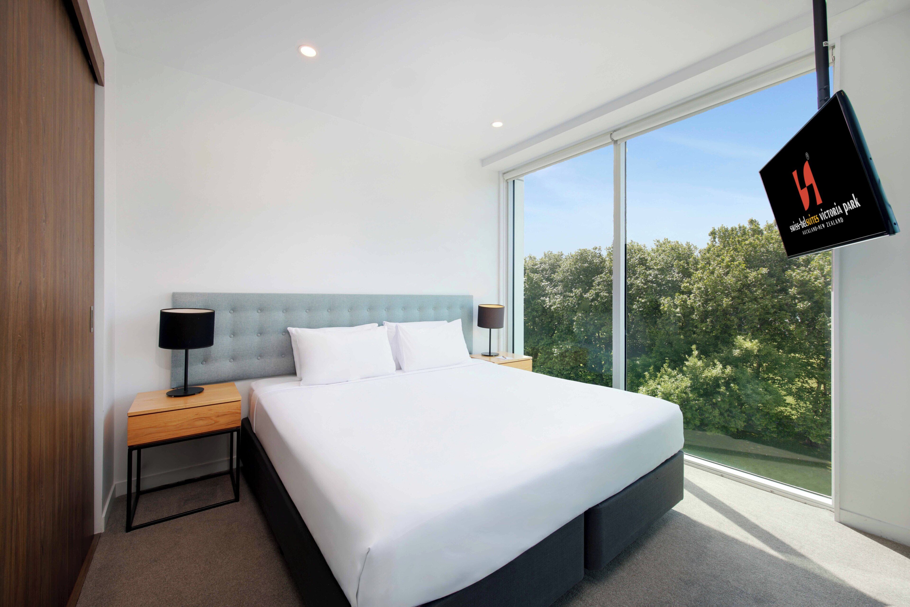 A modern hotel room with a large bed, two bedside tables, a flat-screen TV, and a large window offering a view of trees.