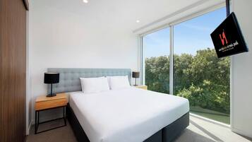 A modern hotel room with a large bed, two bedside tables, a flat-screen TV, and a large window offering a view of trees.