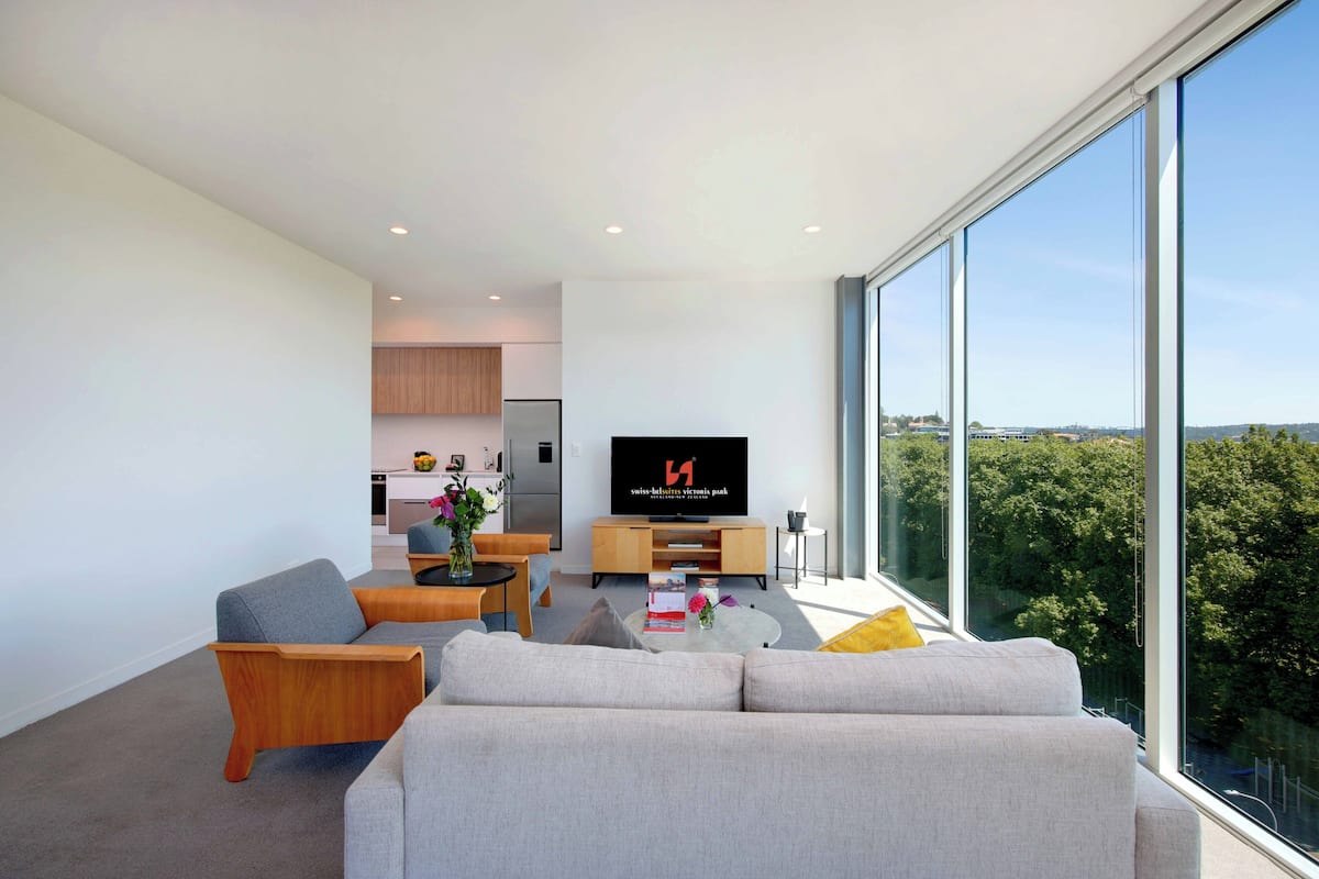 Ein modernes Wohnzimmer mit einer Couch, einem Couchtisch und einem Fernseher. Im Hintergrund ist eine Küche zu sehen und große Fenster bieten Blick auf Bäume.