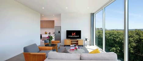 Una sala de estar moderna con un sofá, una mesa de centro y un televisor. Se observa una cocina al fondo y ventanas amplias con vistas a árboles.
