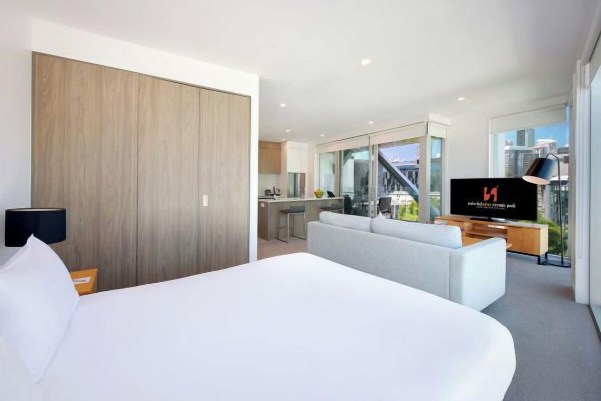 A modern hotel room with a large bed, a sofa, a TV, and a kitchen area visible through a glass partition.