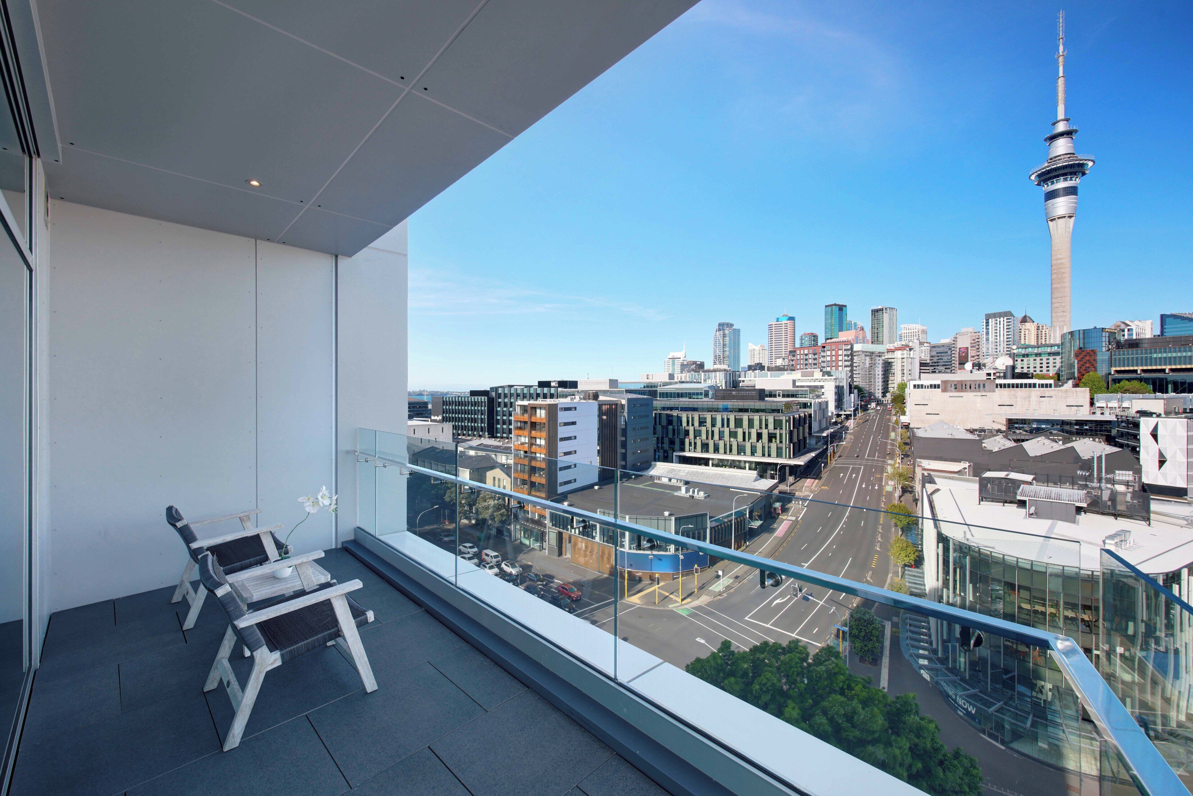 A balcony with two chairs and a table, overlooking a cityscape with modern buildings and a prominent tower.