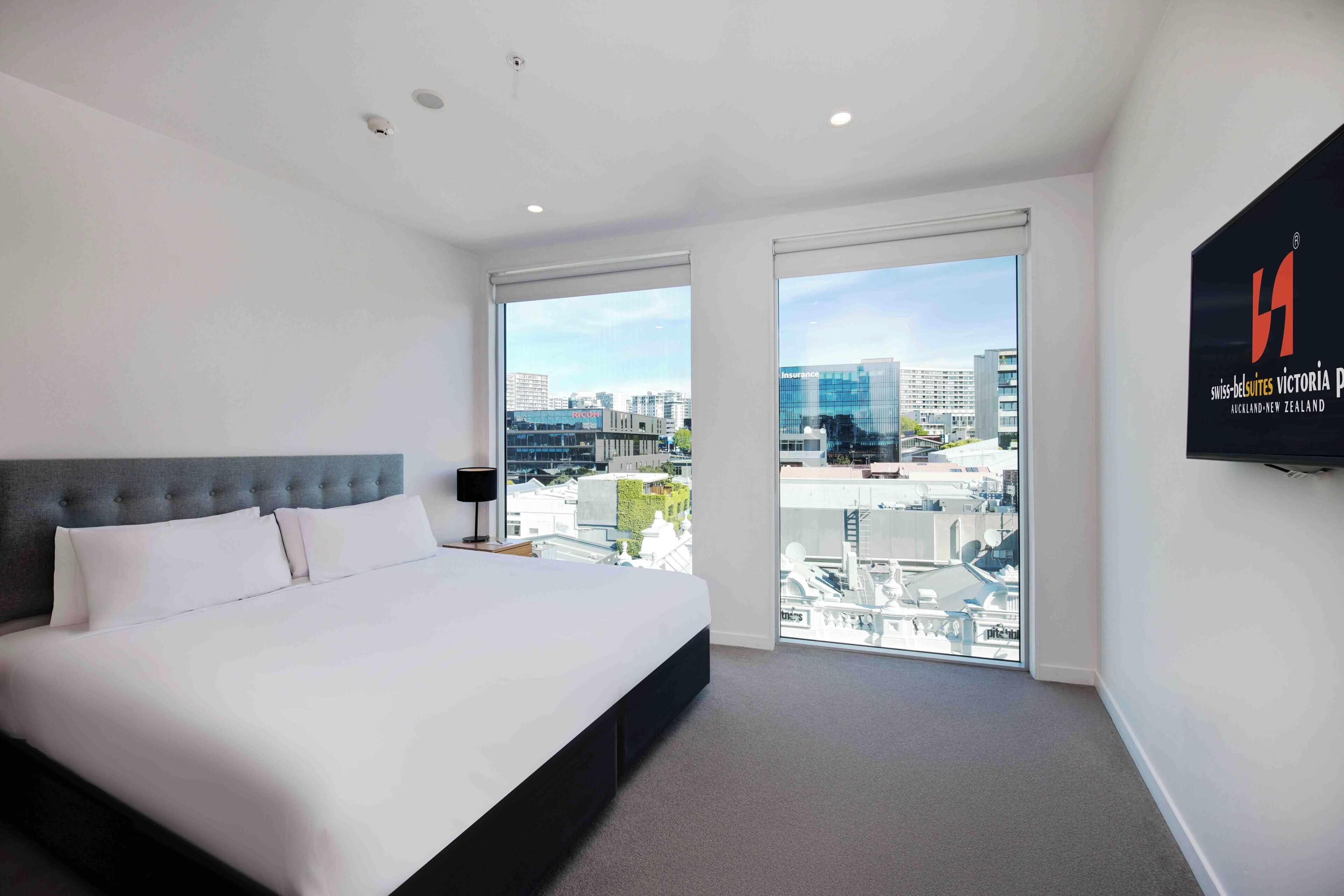 A modern hotel room with a large bed, a city view through floor-to-ceiling windows, and a mounted television on the wall.