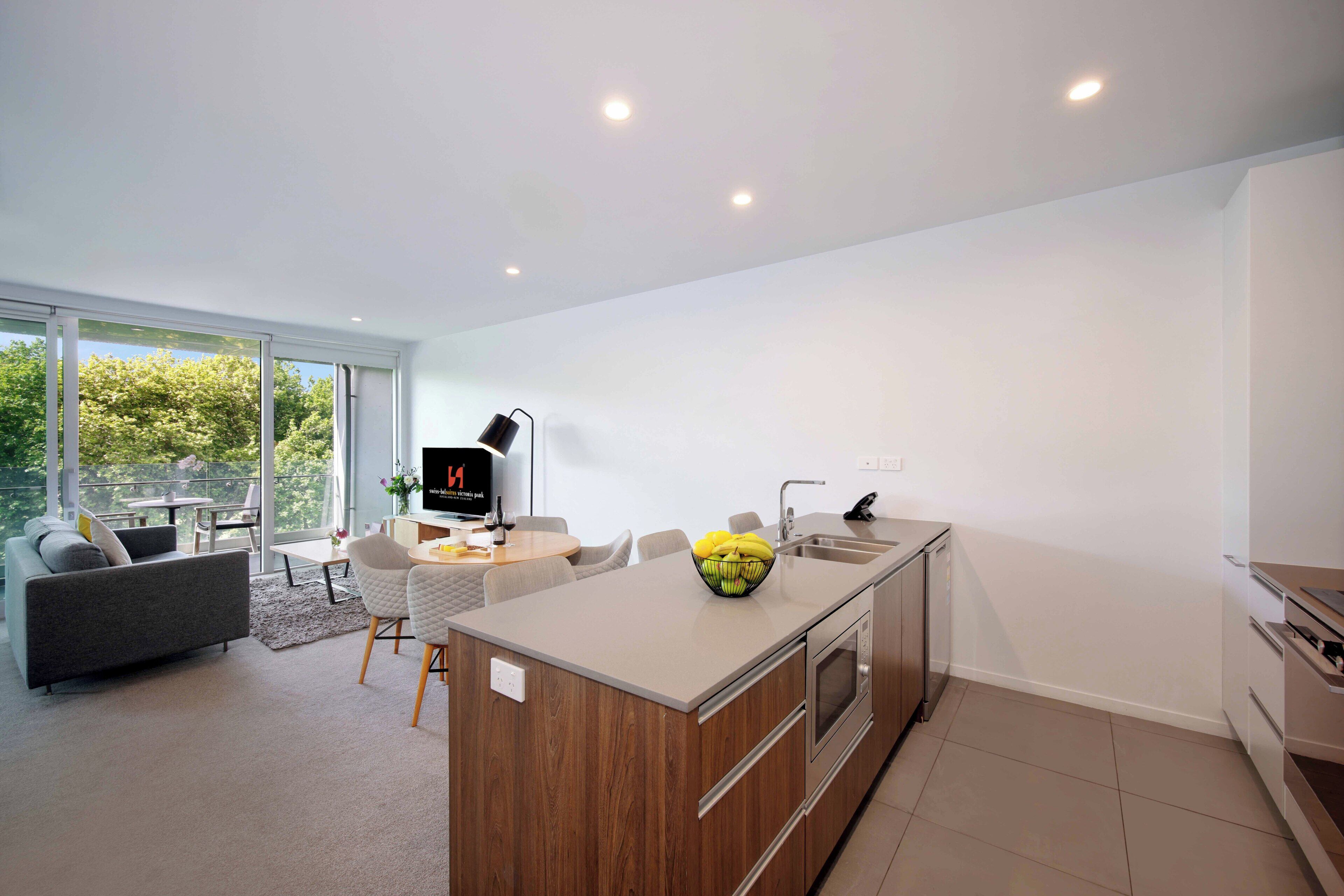 A modern kitchen with a central island, a dining area with a sofa and chairs, and a large window offering a view of greenery.