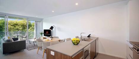 A modern kitchen with a central island, a dining area with a sofa and chairs, and a large window offering a view of greenery.
