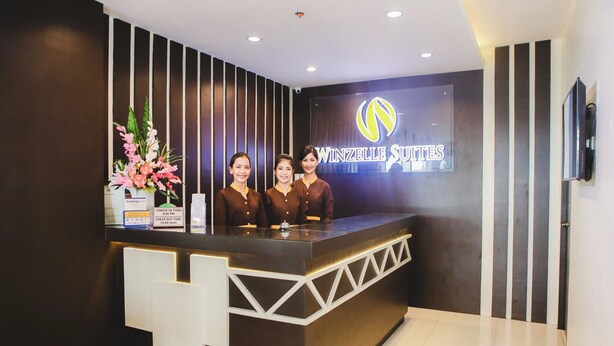 A reception desk with three staff members, a sign for Winzelle Suites, and a check-in/out time display. - Winzelle Suites (Zamboanga)