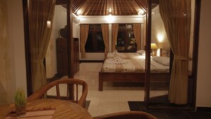 A hotel room with a large bed, a wooden chair, a small table with a plant, and a view of the room through a glass door. - Bucu Beji Ubud (Ubud)