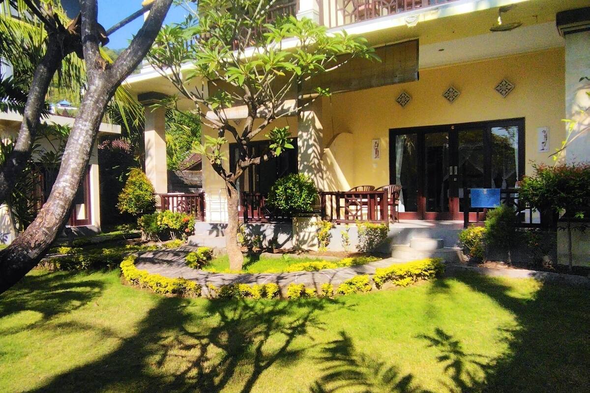 A two-story building with a garden in front, featuring a tree, a small fountain, and a seating area.
