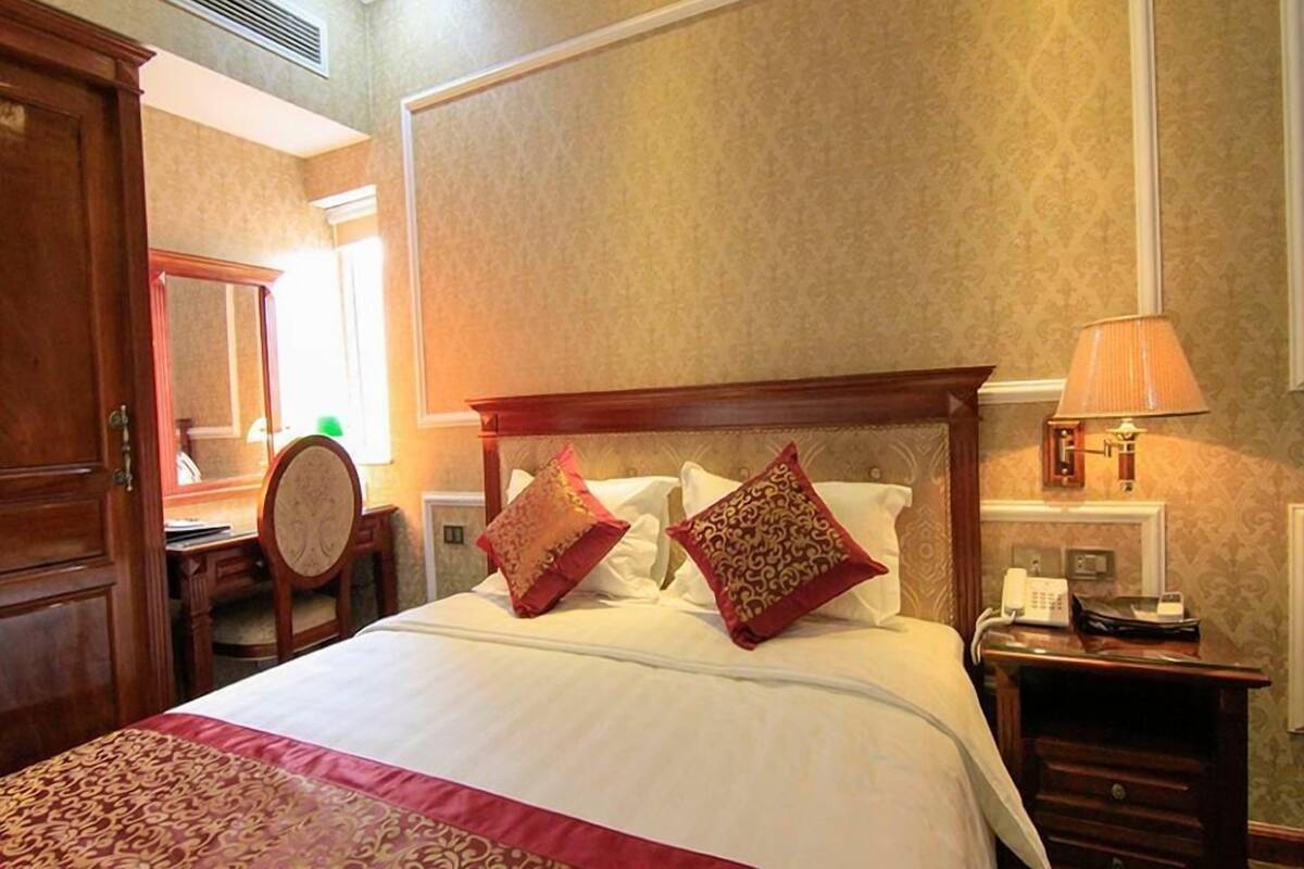 Habitación de hotel con una cama grande, dos almohadones decorativos, una lámpara de noche, una mesita de madera y un espejo.