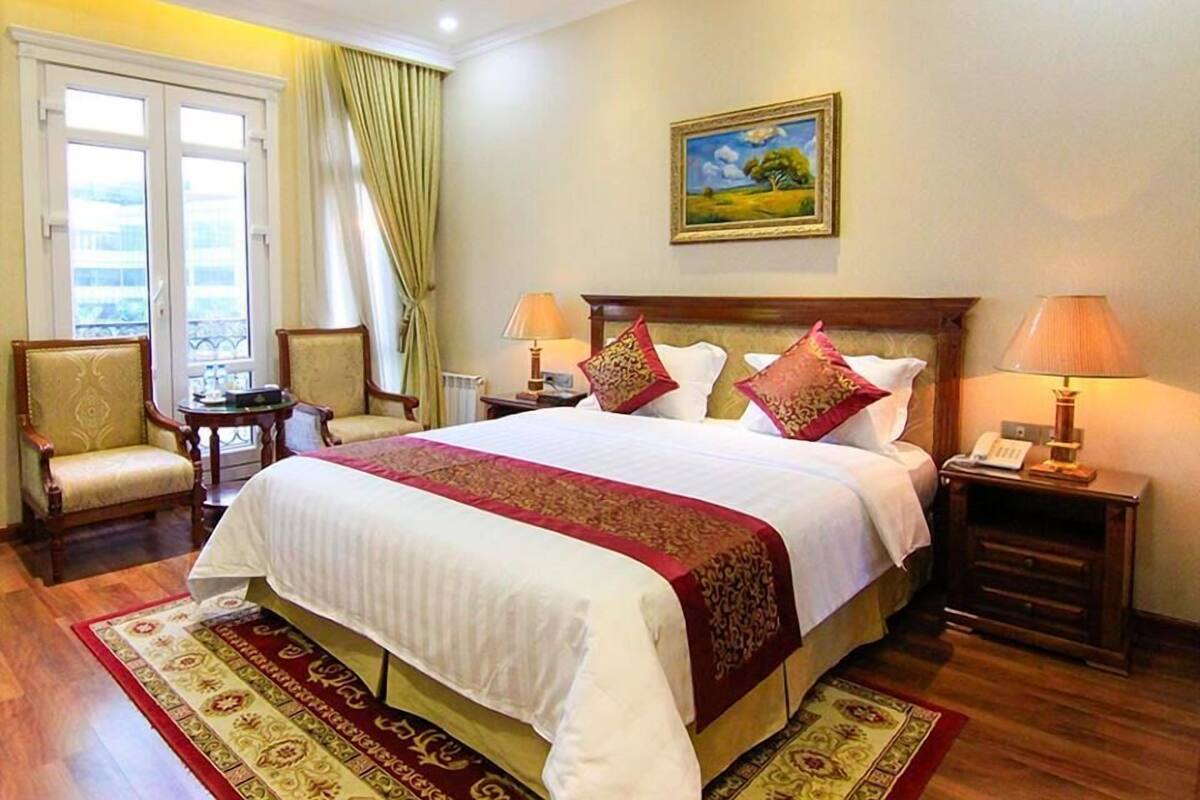 Habitación de hotel con una cama grande, dos mesitas de noche con lámparas, una silla, una mesita pequeña y un cuadro en la pared.