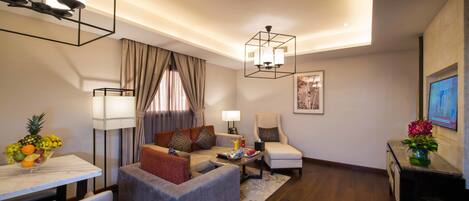 A modern living room with a sofa, armchair, and a coffee table. There is a dining table with a fruit bowl. The room has a ceiling light and a wall-mounted TV.