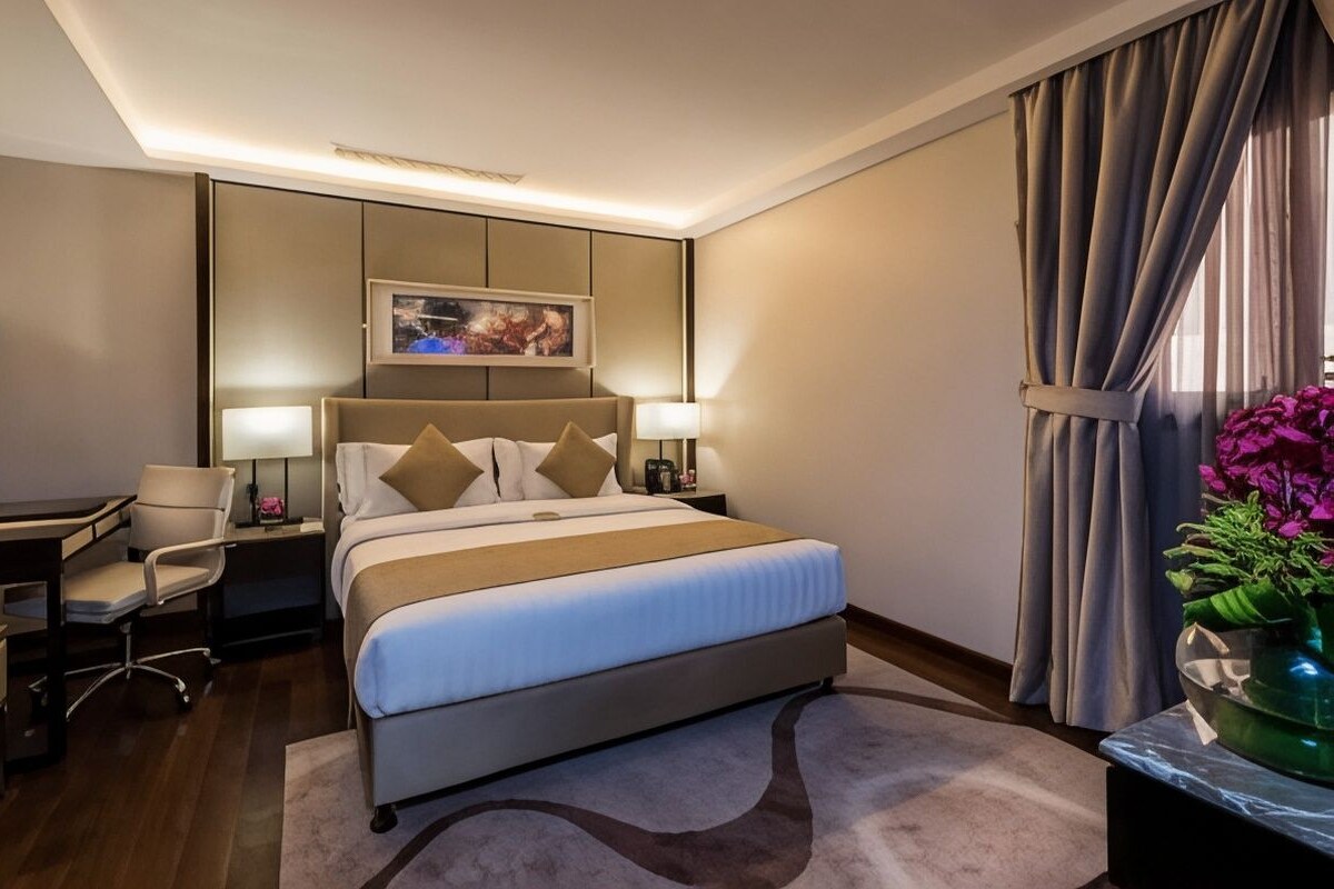 A hotel room with a bed, bedside lamps, a desk, a chair, a large mirror, and a window with curtains.