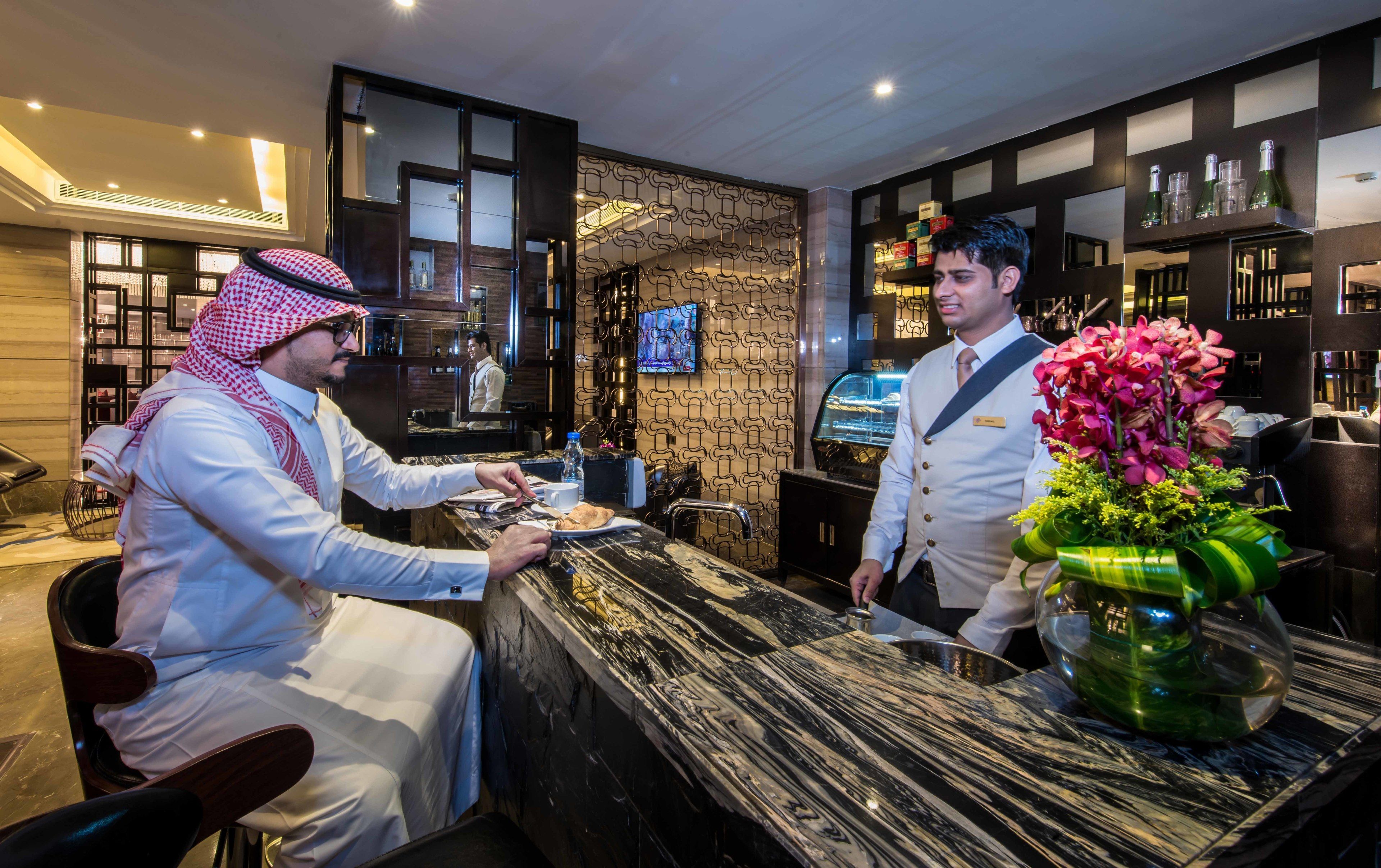 A hotel reception area with a marble counter, a television mounted on the wall, and a bar area with shelves of bottles.
