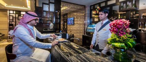 A hotel reception area with a marble counter, a television mounted on the wall, and a bar area with shelves of bottles.