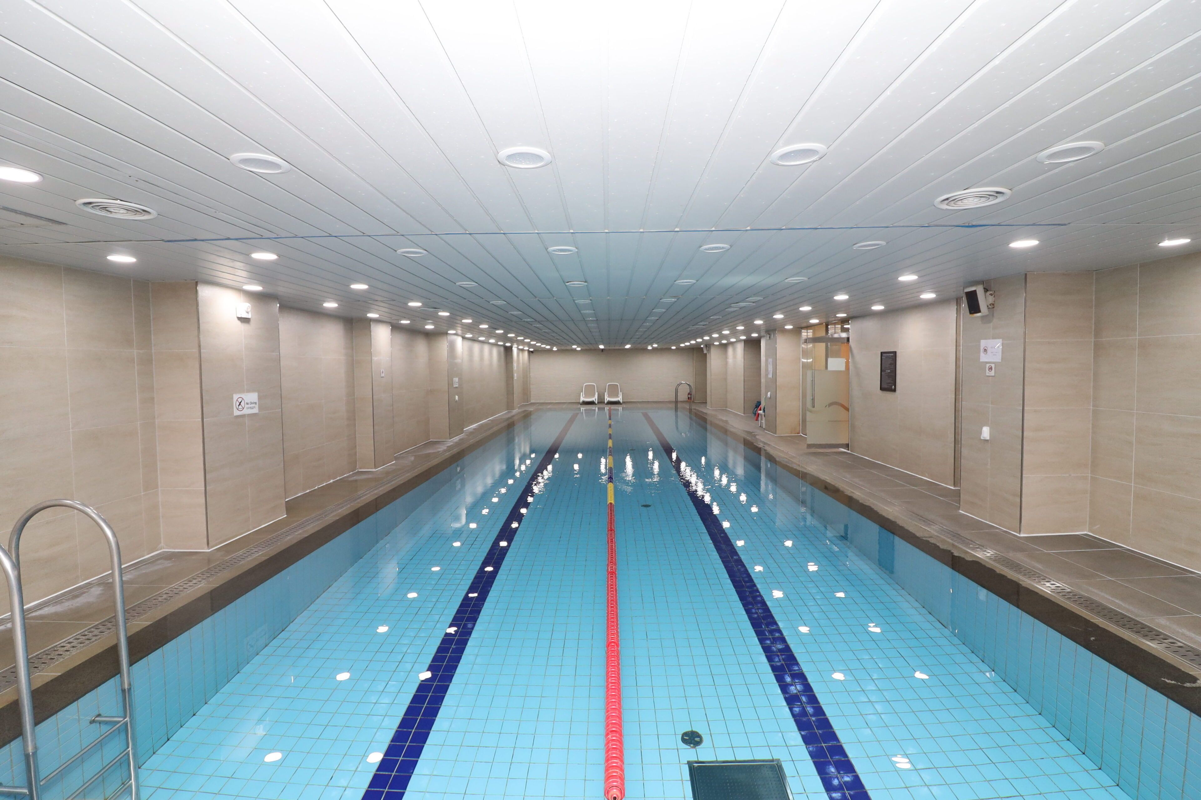 Ein langer, überdachter Schwimmbereich mit gefliesten Wänden und Decke, ausgestattet mit mehreren Lampen und einer zentralen Schwimmbahnteiler.