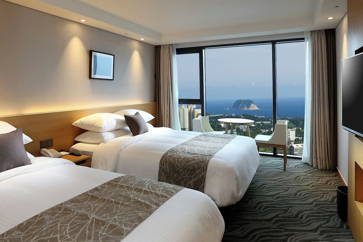 A hotel room with two beds, a desk, a TV, and a large window with a city view.