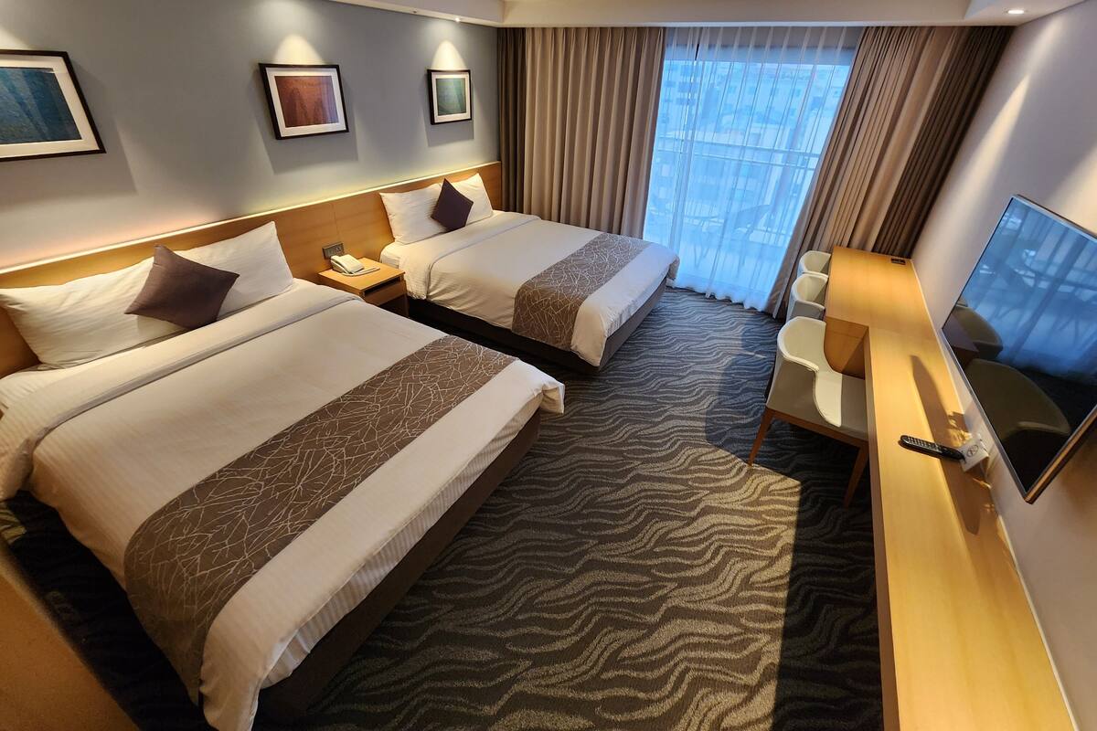 A hotel room with two beds, a desk, a TV, and a window with curtains.