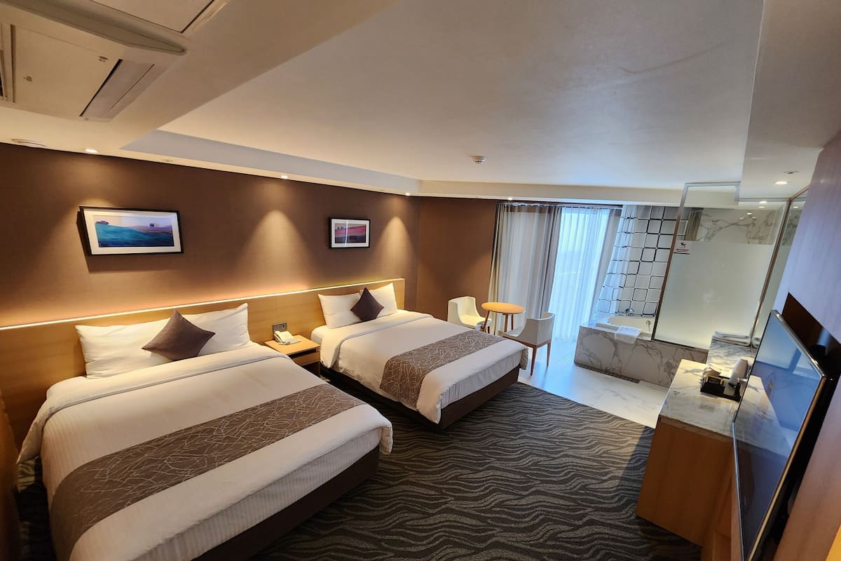 A hotel room with two beds, a desk, a chair, a small table, a mirror, and a bathroom with a glass shower.