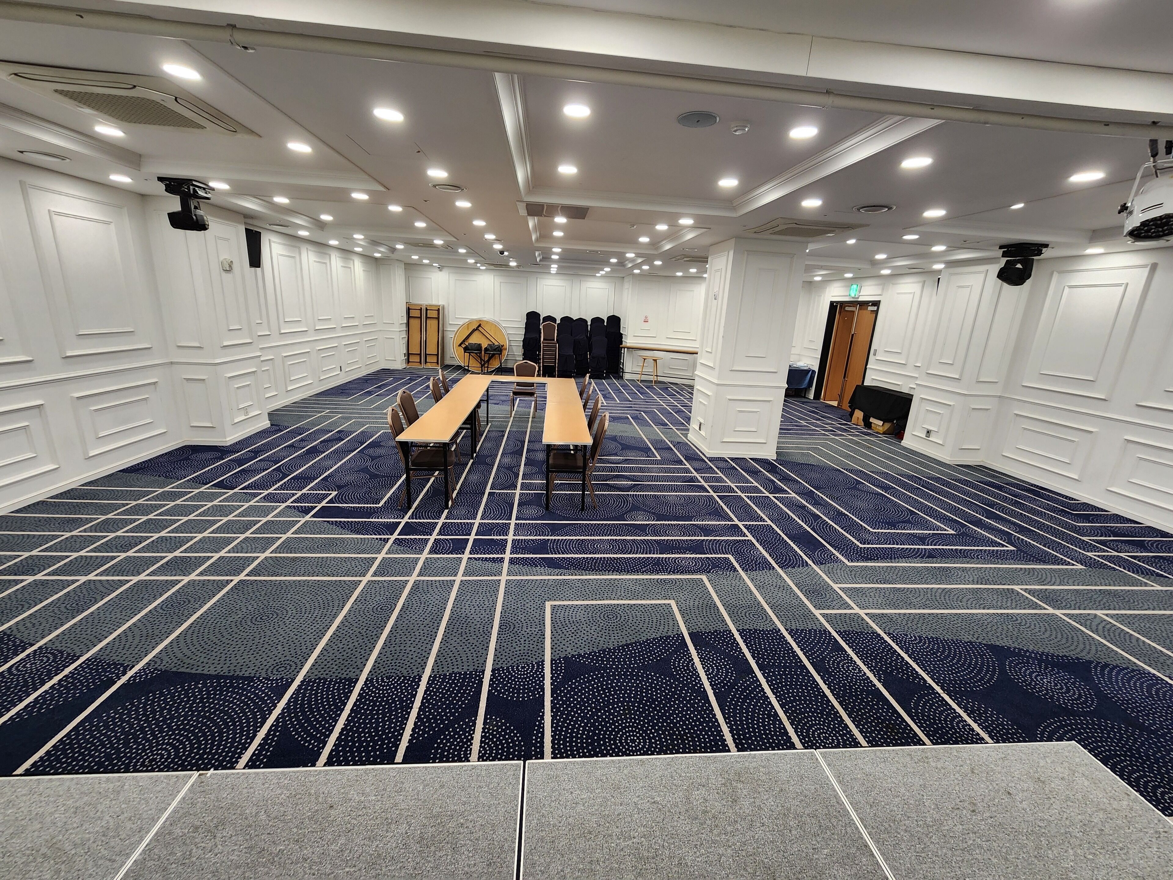 Uma sala de conferência com carpete estampado, paredes brancas e várias mesas e cadeiras.