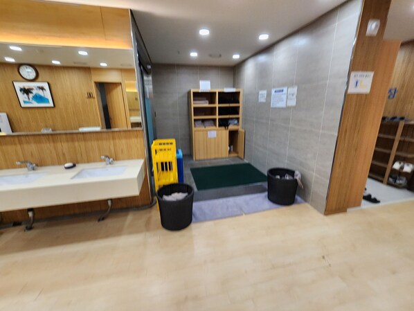 A modern bathroom with a wooden vanity, a sink, a mirror, and a small green mat. There are two black trash bins and a yellow cart.