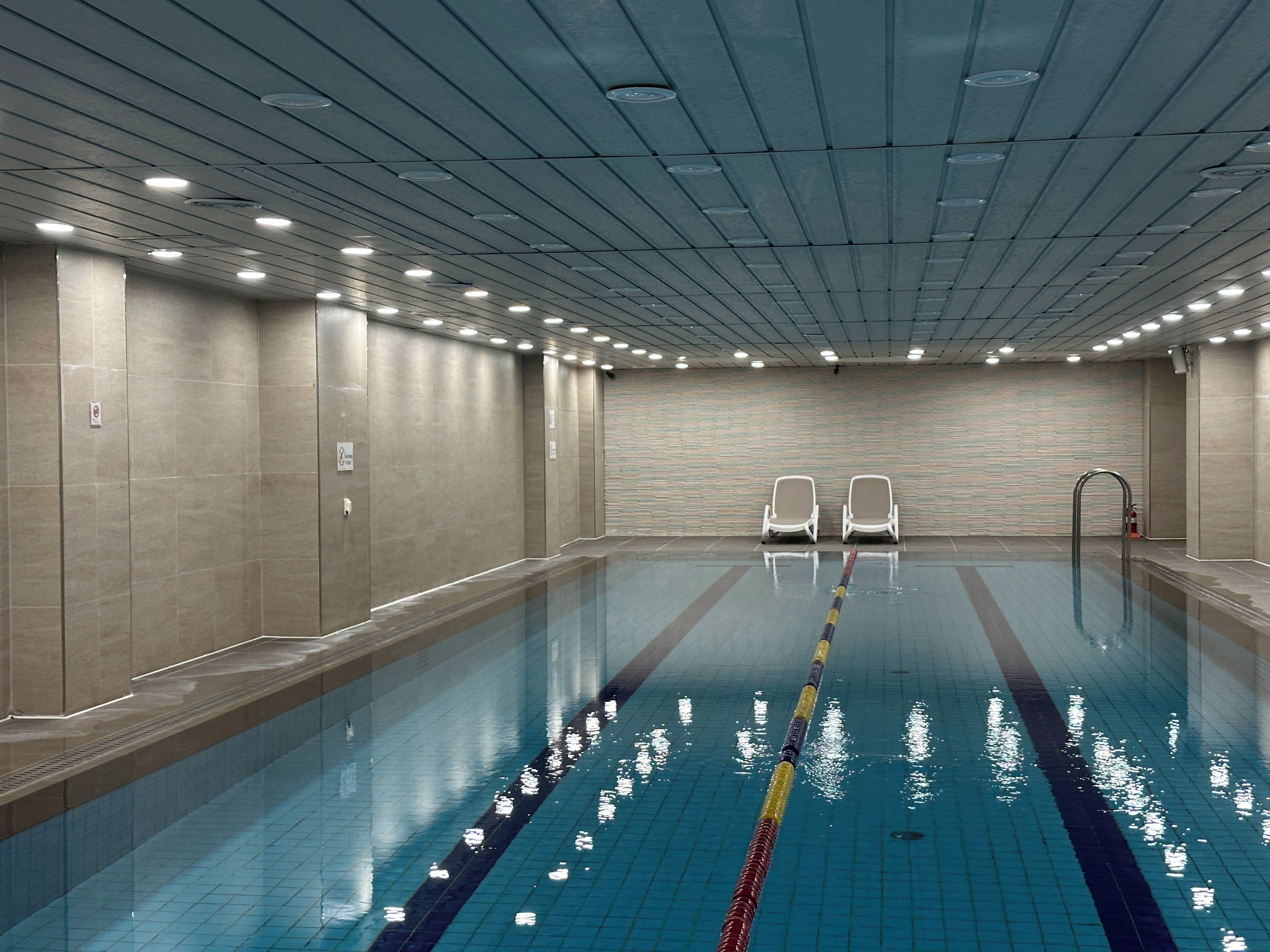 Une piscine intérieure aux murs carrelés, dotée de luminaires au plafond et de deux chaises longues vides.
