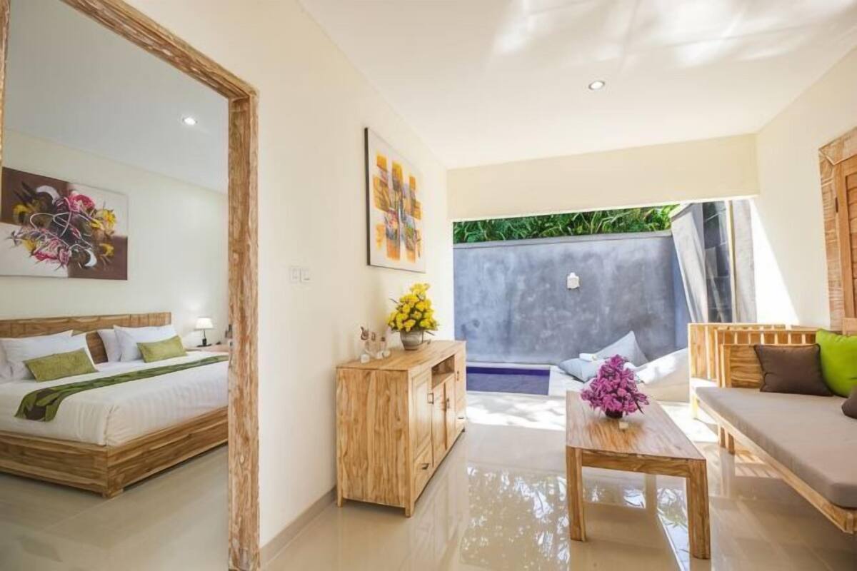 A modern hotel room with a bed, a wooden side table, a sofa, and a view of a blue pool.