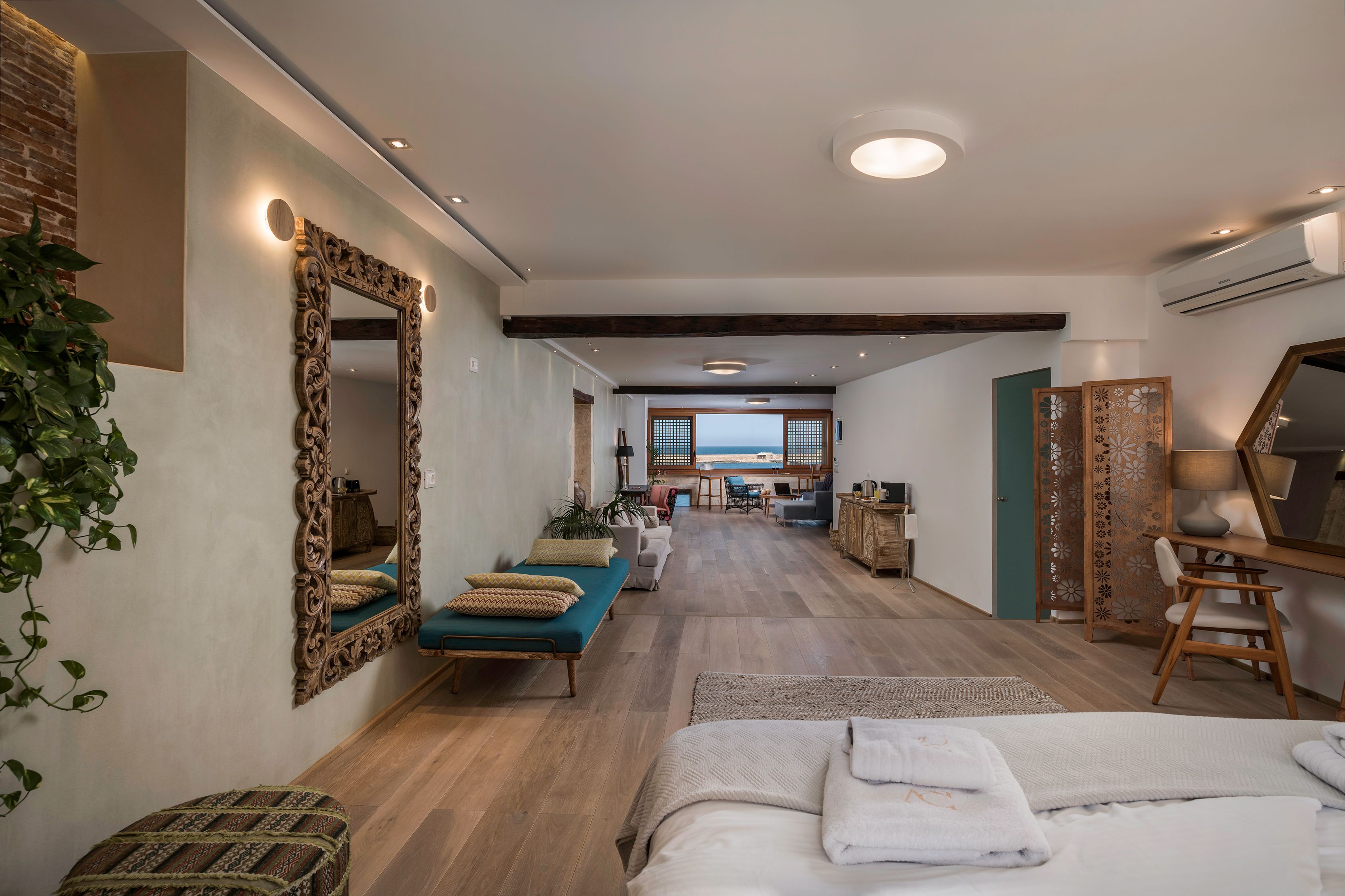 Una habitación de hotel moderna con piso de madera, un espejo grande, una cama con ropa blanca, un sofá azul y un escritorio con una silla.
